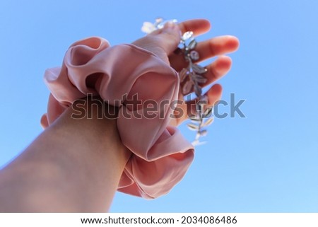 A hand holding floral headband, with a pink scrunchie on a wrist. Clear blue sky in the background.