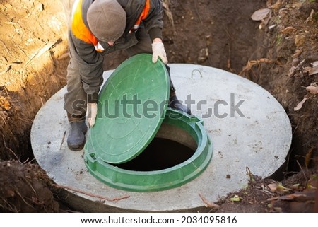 A worker installs a sewer manhole on a septic tank made of concrete rings. Construction of sewerage networks for country houses. Royalty-Free Stock Photo #2034095816