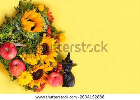 black cat figurine with witch hat, apples and autumn flowers bouquet on yellow background. fall season. symbol of autumn season, thanksgiving, halloween, mabon. flat lay. copy space