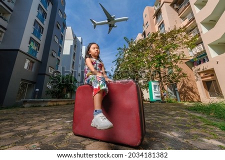 A cute adorable Asian girl about 6-8 years old wearing a vintage dress. An active kid poses for a photo with an old red vintage suitcase alone with a large plane flying in the background.