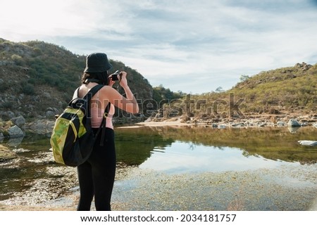 pretty young white hispanic latin tourist girl with backpack and cap standing in front of a lagoon taking pictures of two green mountains and the cloudy sky with her digital camera.