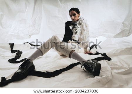 Stylish attractive woman sitting in black hat, jacket and white blouse with flowers. White transparent crumpled paper with black ribbons on background