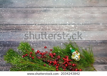 A photo of a snowman bauble with red twigs and pine branch on a wooden background. Copy space. Snow overlay.