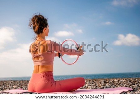 Young woman with black hair, fitness instructor in pink sports leggings and tops, doing pilates on yoga mat with magic pilates ring by the sea on the beach. Female fitness daily yoga concept. Royalty-Free Stock Photo #2034244691