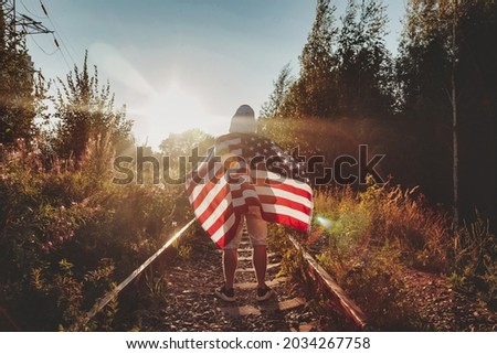 Young man walks along railway tracks of road with an American flag towards sunset. Male stands on an abandoned road with rails in rural area with stripes and stars flag. Concept of travel, freedom