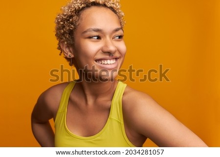 Girl of mixed race without makeup, smiling widely, showing healthy white teeth, wearing sports wear of yellow color, having nose and ears pierced, looking aside. Natural beauty concept