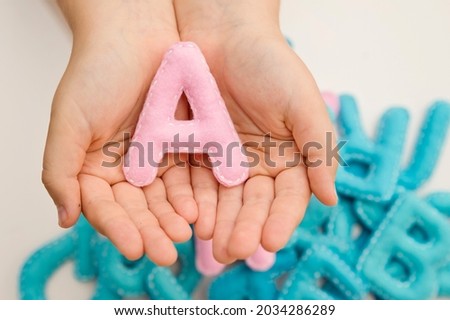 Stuffed felt letter of the alphabet in open hands of child. Kid holding pink color handmade capital letter " A " in background of many other blue color letters. Montessori colors.