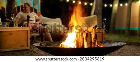Cast iron fire pit campfire place at forest beach camping with brgiht burning flame at evening time against light bulb garland and trees. Friends sit near camp bonfire stove at warm summer night Royalty-Free Stock Photo #2034295619