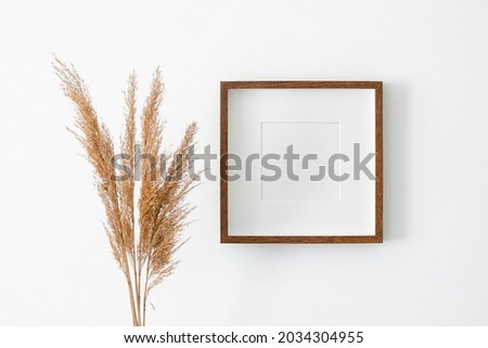 Square wooden frame with passepartout on white wall with dry plant decorations. Blank frame mockup for artwork, print or photo presentation.