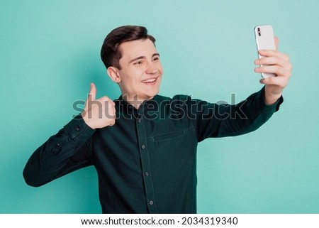Portrait of cheerful positive reliable man hold mobile phone make selfie raise thumb up on teal background