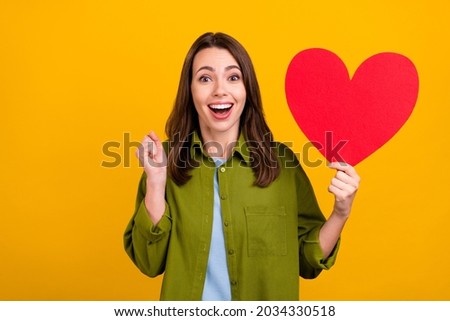 Photo of shiny lucky young woman dressed green shirt smiling holding big red heart rising fist isolated yellow color background
