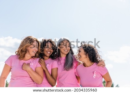 Cheerful multiethnic women with ribbons of breast cancer awareness hugging outdoors Royalty-Free Stock Photo #2034335960