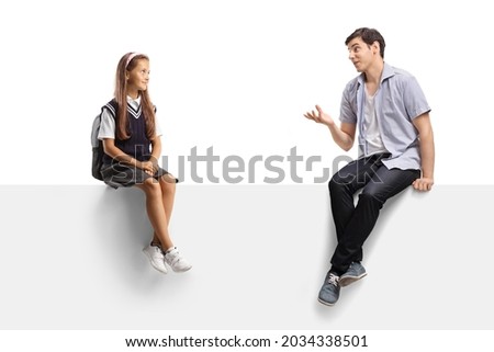 Young man sitting on a blank panel and talking to a schoolgirl isolated on white background