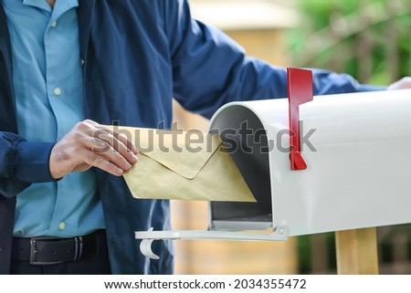 Handsome young postman putting letter in mail box outdoors Royalty-Free Stock Photo #2034355472