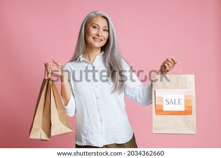 Positive middle aged Asian female model holds paper shopping bags with bright Sale sign standing on pink background in studio