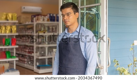 Young male owner is preparing to give warm welcome to customers for his local startup bakery or coffee shop in front of the shop. Smiling attractive owner with glasses stands for welcoming.