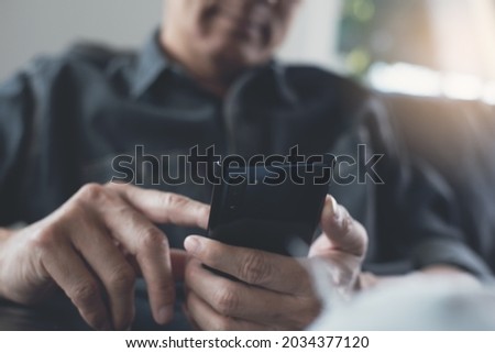 Asian man in casual clothes sitting at comfortable couch holding smartphone surfing social networks, chatting with his friend via social media app at home