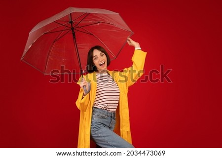 Excited young lady enjoying rainy weather, standing under transparent umbrella and smiling to camera over red studio background, copy space