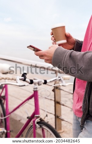 vertical photo of an unrecognizable young man with a drink using a mobile phone next to a vintage bike, concept of technology of communication and urban lifestyle, copy space for text