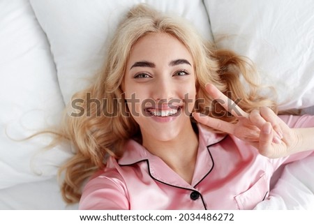 Happy, relaxed after night sleep. Sweet dreams, good morning, new day, weekend, holiday. Glad young european lady in pink pajama show peace sign, takes photo on white comfort bed with pillow, close up