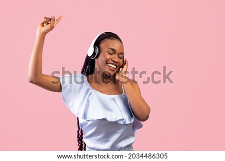 Positive black woman in wireless headphones dancing to popular music on pink studio background. Carefree African American lady clubbing to favorite song, enjoying cool playlist