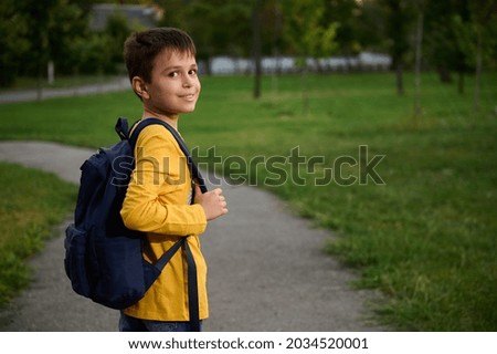 Side view of a schoolboy wearing yellow sweatshirt with school bag backpack walking on the path in public park, going home after school, cute smiling to the camera