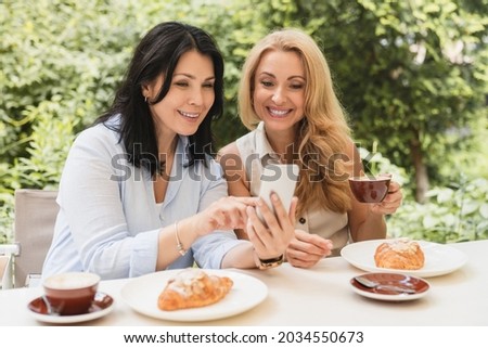 Two caucasian businesswomen sisters best friends girlfriends showing news social media photos on smart phone cellphone sitting in cafe and drinking coffee during breakfast.
