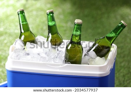 Blue plastic cool box with bottles of beer and ice cubes on blurred green background