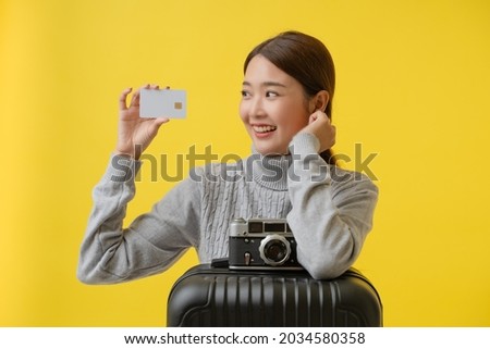 Portrait of female Asian tourist, dressed in grey turtleneck sweater showing credit card with a retro camera and black luggage on a yellow background. 