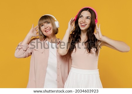 Two young smiling happy daughter mother together couple women wearing casual beige clothes headphones listen to music isolated on plain yellow color background studio portrait Family lifestyle concept