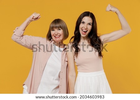 Two young strong sporty fitness daughter mother together couple women in casual beige clothes show biceps muscles on hand demonstrating strength power isolated on plain yellow color background studio Royalty-Free Stock Photo #2034603983