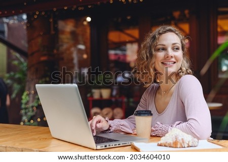 Young smiling happy woman in casual clothes sit alone at table in cafe shop eat breakfast croissant hold use work on laptop pc computer drink coffee relaxing in restaurant during free time indoors. Royalty-Free Stock Photo #2034604010
