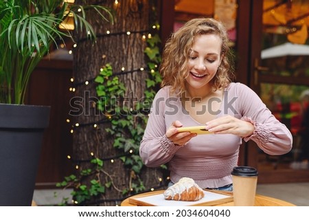 Young smiling happy woman 20s in casual clothes sitting alone at table in coffee shop cafe eat breakfast taking photo of lunch meal by mobile cell phone relaxing in restaurant during free time indoors