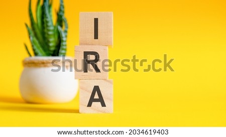 Three wooden cubes with letters - IRA on yellow table, space for text in right. Front view concepts, flower in the background. IRA - short for individual retirement account