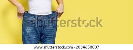 The concept of diet, proper nutrition, weight loss. Slim Woman Showing Loose Jeans and her Loss Weight. Woman white t-shirt and oversize jeans isolated on yellow background. Banner with copy space.