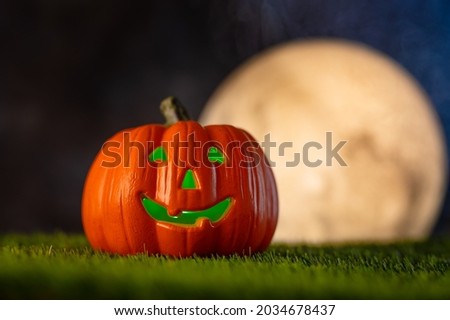 An orange pumpkin in the form of a head with a smile on green grass against the background of a dark night sky and a large white moon. Halloween holiday. Postcard, banner, poster.