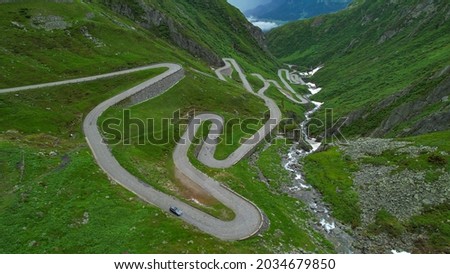 DRONE: Tourists on road trip across the mountains of Switzerland drive along hairpin turns of Gotthardpass. Grey colored tourist car cruises along the scenic switchback road of Passo San Gottardo. Royalty-Free Stock Photo #2034679850
