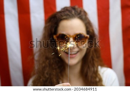 Blurred Young redhead Hispanic and Caucasian woman holding a sparkler in focus standing in front of the American flag.