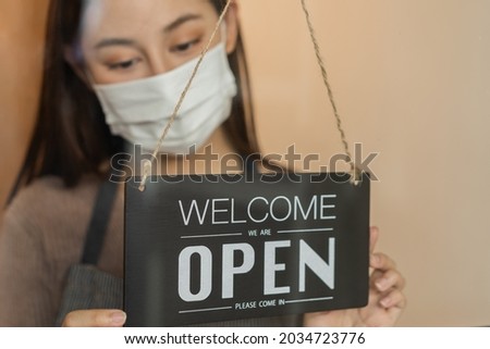 Smiling asian young owner retail,coffee shop woman turning sign board to open wearing face mask,protect to pandemic of coronavirus,reopen store after close lockdown quarantine in covid to new normal.