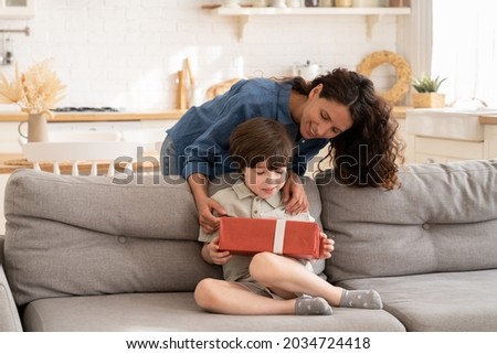 Birthday of child in family: happy mother giving gift in decorated present box to excited kid on anninersary or son day. Caring mom prepare surpise to little preschool boy sit cheerful on sofa at home Royalty-Free Stock Photo #2034724418