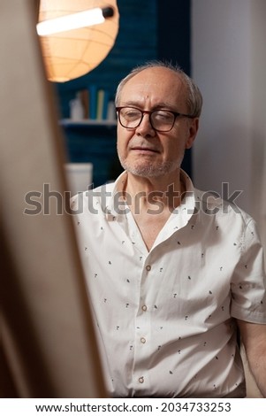 Portrait of senior artist looking at canvas with masterpiece sitting in workshop at home. Close up of elderly man with artistic hobby thinking about drawing with professional instruments