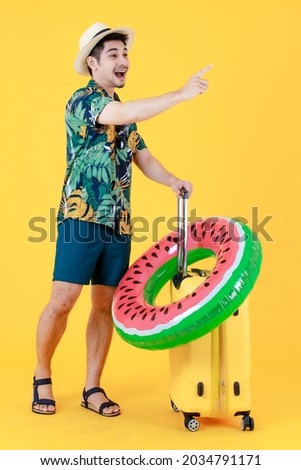 Profile shot, young Asian man in colorful Hawaiian shirt gives index finger up and pull yellow suitcase. Full body studio portrait on yellow background. Happy Summer holiday travel concept