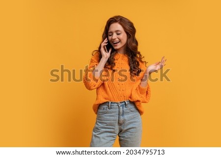 Attractive red head woman in stylish fall outfitm sweater and jeans posing over  yellow   background in studio.  Talking by  smartphone with suprice face. 