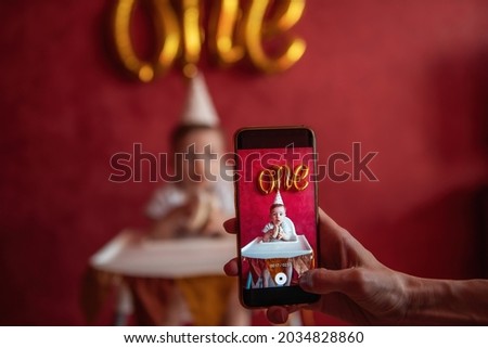 Mothers hand takes pictures of son on a smartphone. A little boy of one year in festive hat holding birthday cake with a candle in hands. On red plain background gold foil balloons One. Social Media
