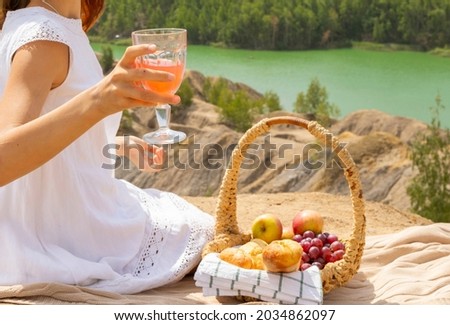 teenage girl in white dress is sitting on edge of cliff next to picnic basket and having fun during lunch, background of stunningly beautiful landscape with rocks and lake outdoor