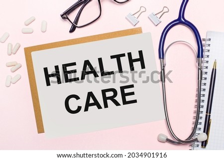 On a light pink background, a stethoscope, white pills and clips for paper, glasses in black frames and a sheet of paper with the text HEALTH CARE. View from above. Medical concept
