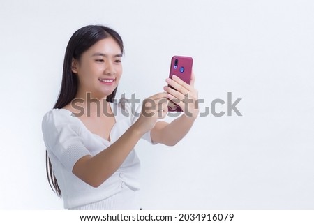 Portrait of Asian beautiful girl who has black long hair in white shirt, is holding the smartphone in her hand and smiling. She take a selfies photo on white background.