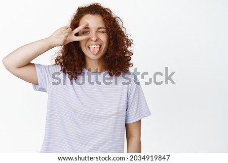 Positive young woman in t-shirt showing peace sign, winking and sticking tongue carefree, silly happy face, standing over white background.