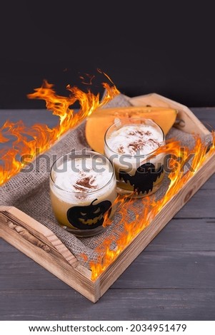Two glass glasses with Halloween-themed drawings with pumpkin latte on a wooden tray with a flame.
