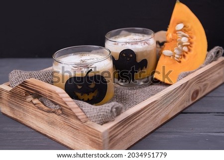 Close-up of glass glasses with Halloween-themed drawings with a pumpkin latte and a piece of pumpkin on a wooden tray .